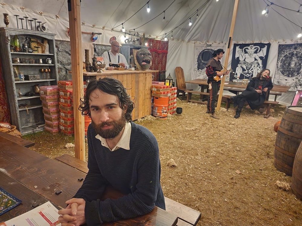 Owen sitting at a wooden table inside a medieval fantasy LARP tent with actors in costume in the background