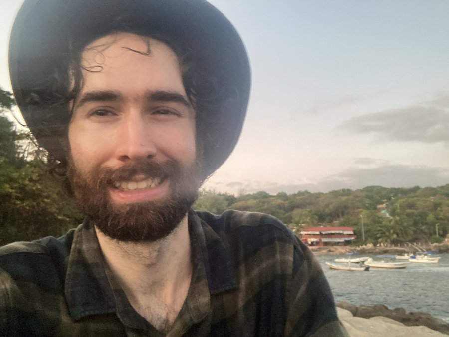 Owen smiling in a selfie on a tropical beach in Mexico with boats in the background
